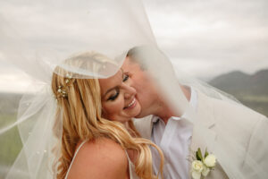 A bride and groom portrait. The groom is kissing the brides cheek. They are under the wedding vail. Captured by Michelle Betz Photography