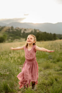 A little girl with arms out flying like an airplace. Surrounded by Denver Colorado mountains and green grass. Captured by Colorado Springs family photgrapher, Michelle