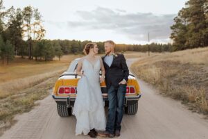 Bride and groom, leaning on an old sports car. They are smiling at each other. 