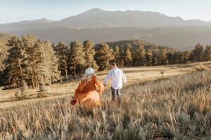 A couple running down a hill holding hands. With Colorado mountains in the background, and fall grass. Captured by Michelle Betz Photography