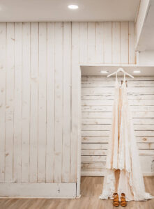 A detail shot of the wedding dress hanging in a door frame, with wedding shoes. Shiplap covered walls.