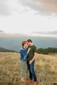 A couple leaning on each other with thier forheads together. Surrounded. by green grass and Colorado Springs mountains in the distance.