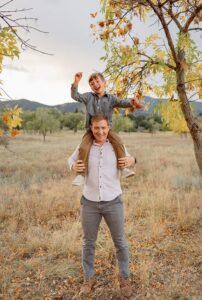 A dad and son portrait. The son is sitting on his dads shoulders and laughing. In the fall, in Colorado Springs.