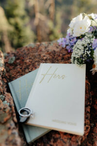 A detail photo of his and her vow books, bouquet, and rings. Photo taken at a Evergreen, CO wedding venue.