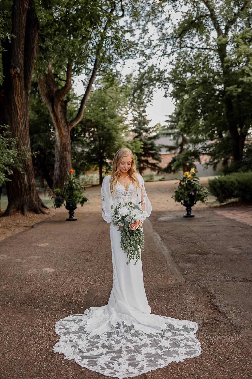 A bridal portrait. She is looking down at her bouquet. Beautiful whtie wedding dress, surrounded by trees.