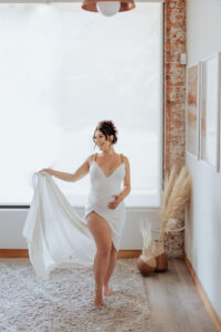 maternity photo of a women in a white dress, holding up the hem of her dress, hand on her stomach, smiling to the side.