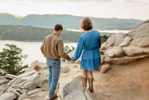 A couple holding hands and walking together for their engagement photos. Surrounded by a lake and mountains. 