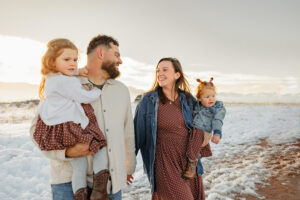 A family holding hands and smiling at each other. Wintertime with snow on the ground, at sunset. Captured by Michelle, a family photographer