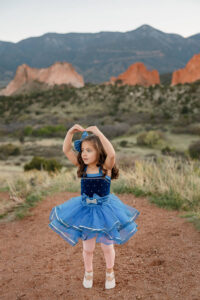 A little girl in her dance outfit, holding her hands up like a ballerina. Surrounded by red rocks and mountains. 