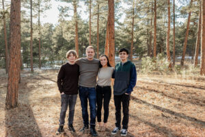 A family of 4, smiling at the camera for a portrait. Photo taken at Fox Run Park in the woods. 